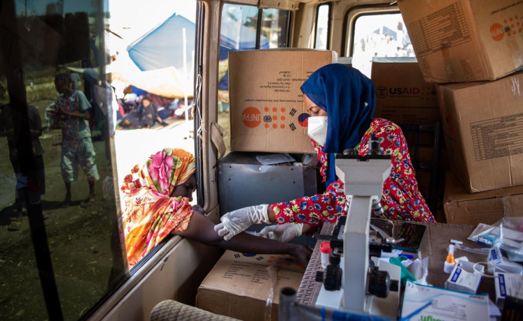 A masked and veiled woman sitting in a van behind a microscope and in front of UNFPA-labelled boxes administers medication to woman's arm as she leans in through the open window.